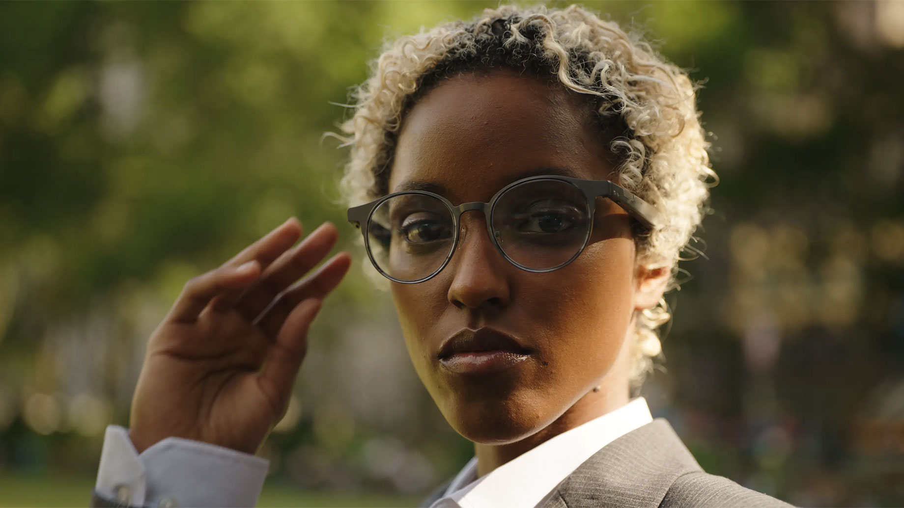 Person with short blond curls wearing round dark-framed smart glasses and a gray blazer outdoors, with one hand raised beside the frames.