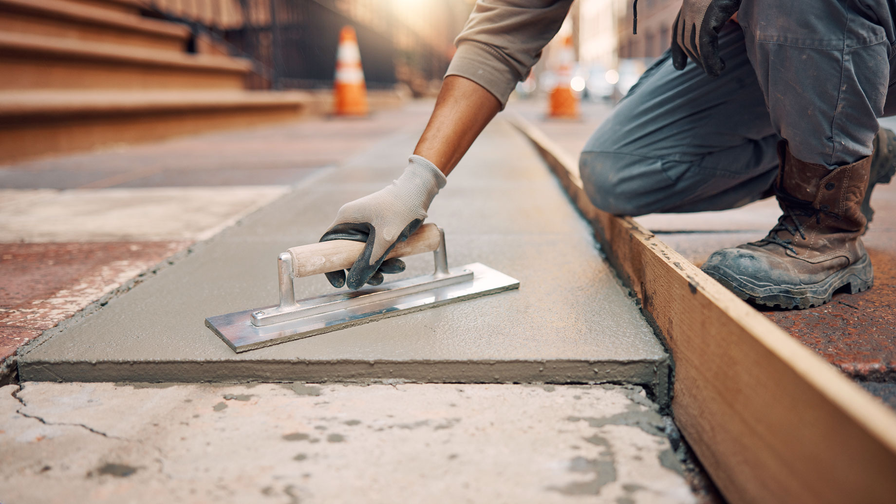 Gloved contractor smoothing fresh sidewalk concrete with a hand trowel beside wooden formwork and brownstone steps.