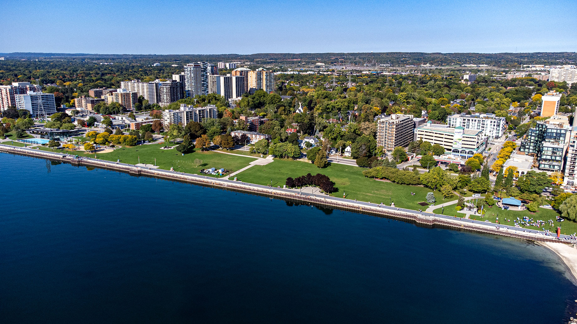 Aerial view of Spencer Smith Park in Burlington, showing green lawns, a waterfront promenade, mature trees, and downtown buildings beside Lake Ontario.