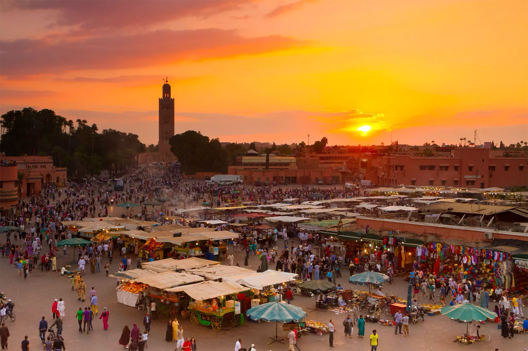 Crowds and market stalls fill Marrakech’s Jemaa el-Fnaa beneath an orange sunset, with the Koutoubia minaret rising in the distance.