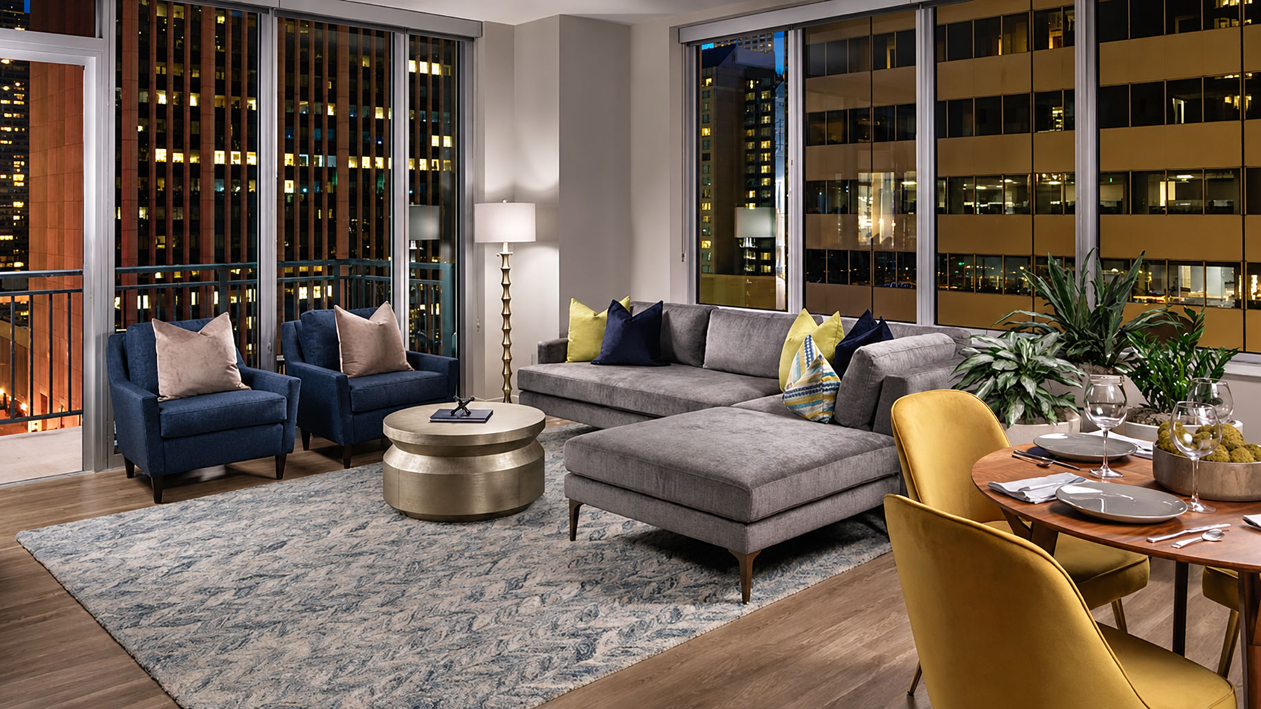 Gray sectional, navy armchairs and a dining table beside floor-to-ceiling windows with nighttime city views at The Quincy.