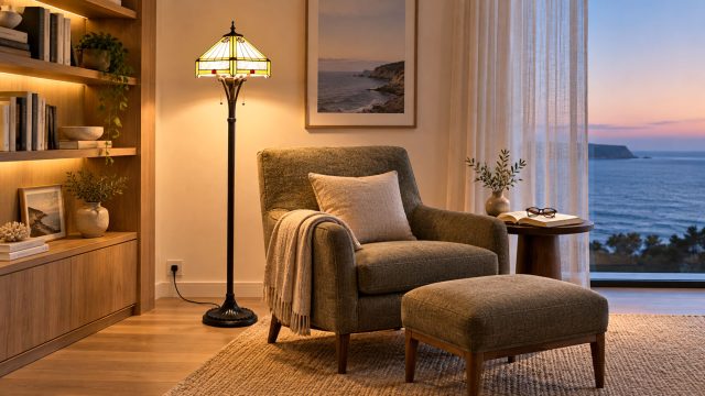 Tiffany-style floor lamp beside an armchair and ottoman, with illuminated bookshelves and an ocean view at dusk.