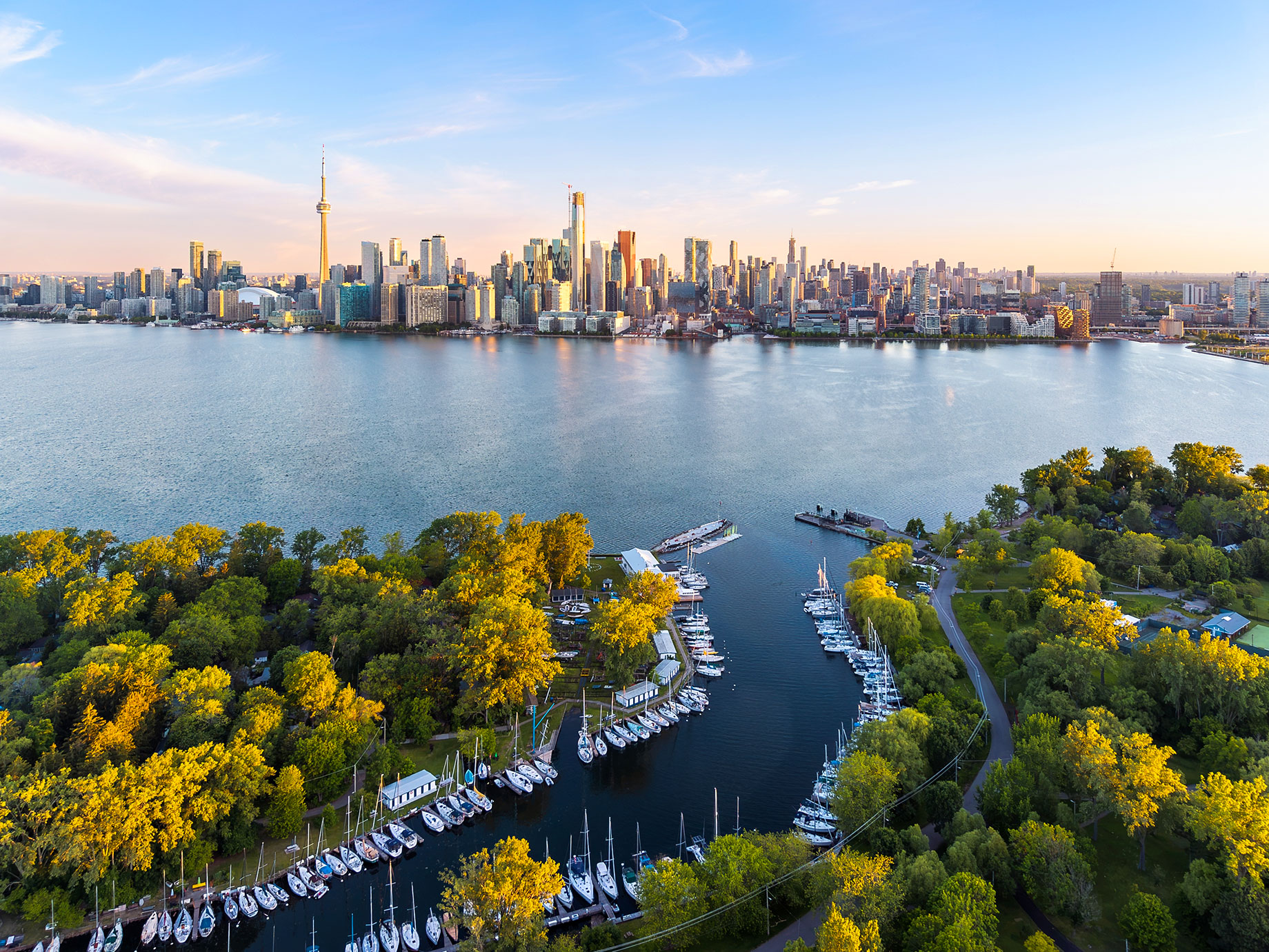 Aerial view of Toronto’s skyline and CN Tower across the water, with tree-lined island shores and moored sailboats in the foreground.