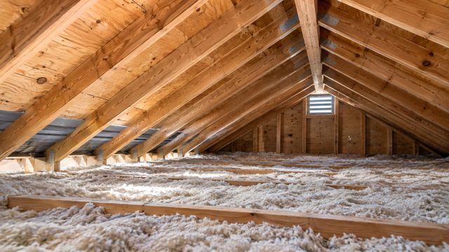 Unfinished attic with exposed wooden rafters, loose-fill insulation, ventilation baffles near the eaves, and a louvered gable vent.