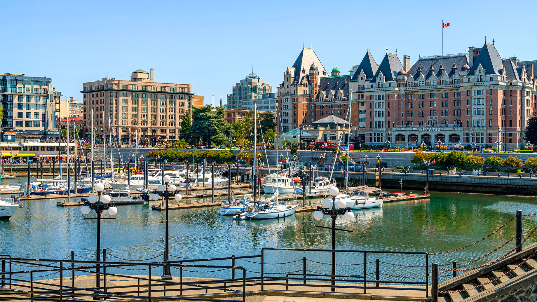 Sailboats moored in Victoria’s Inner Harbour, with the Empress Hotel, waterfront buildings, and a promenade behind them.