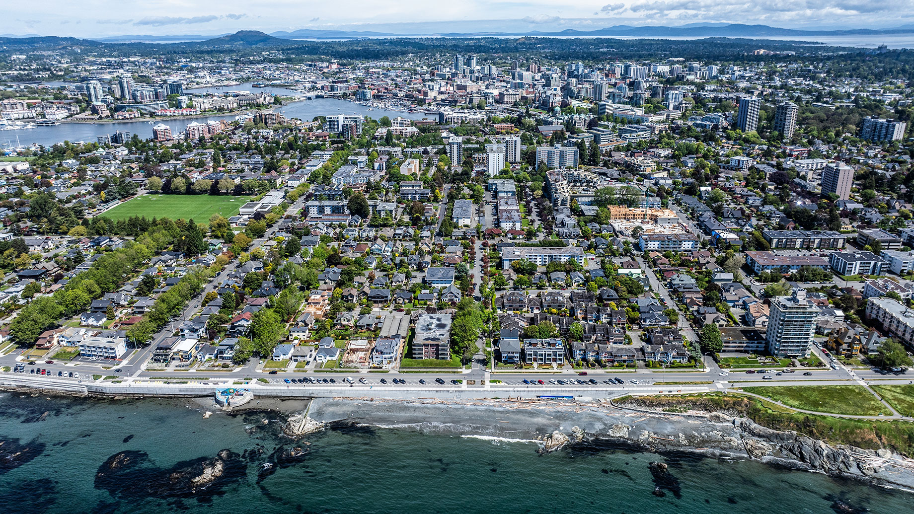 Aerial view of Victoria’s James Bay neighbourhood, showing a rocky shoreline, tree-lined residential streets, and downtown buildings around the harbour beyond.