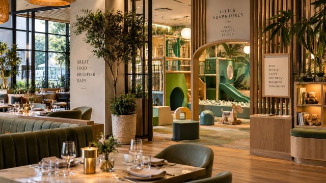 Restaurant tables and green seating beside an indoor play area with a wooden arched entrance, green slide, and ball pit.