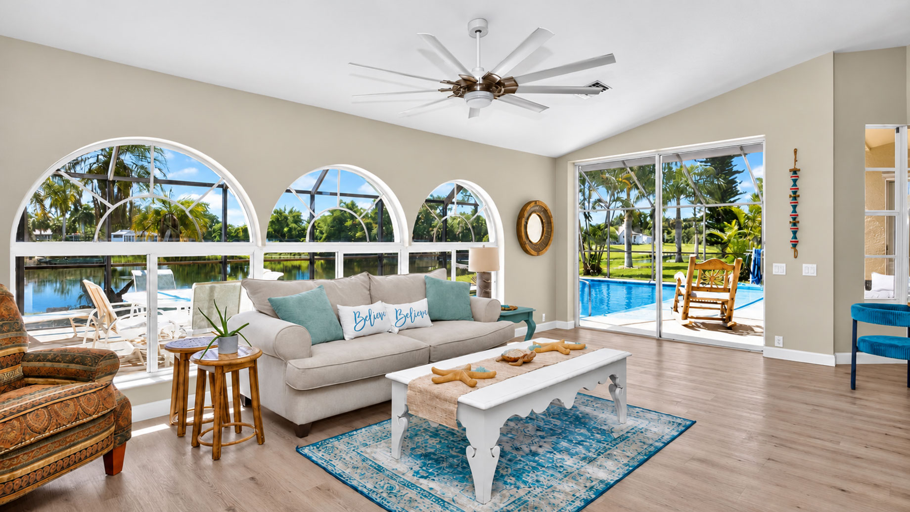 Bright living room with arched windows, a neutral sofa and sliding glass doors overlooking a screened pool and waterfront.